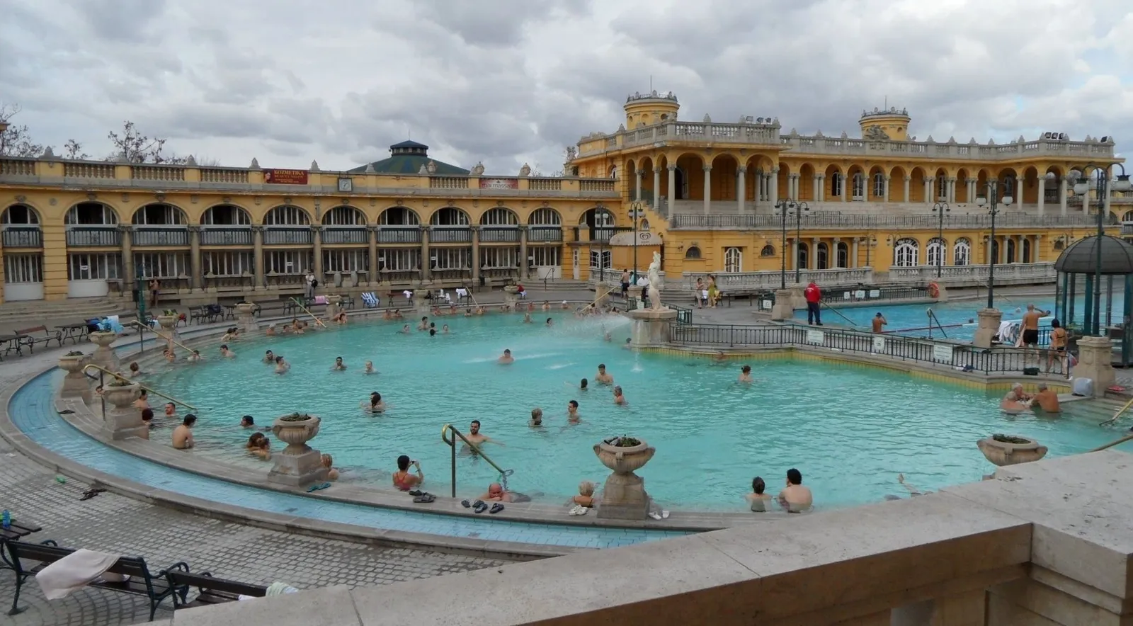 Bathers in Széchenyi's outdoor thermal pool on an overcast day, the arcaded yellow wings around it