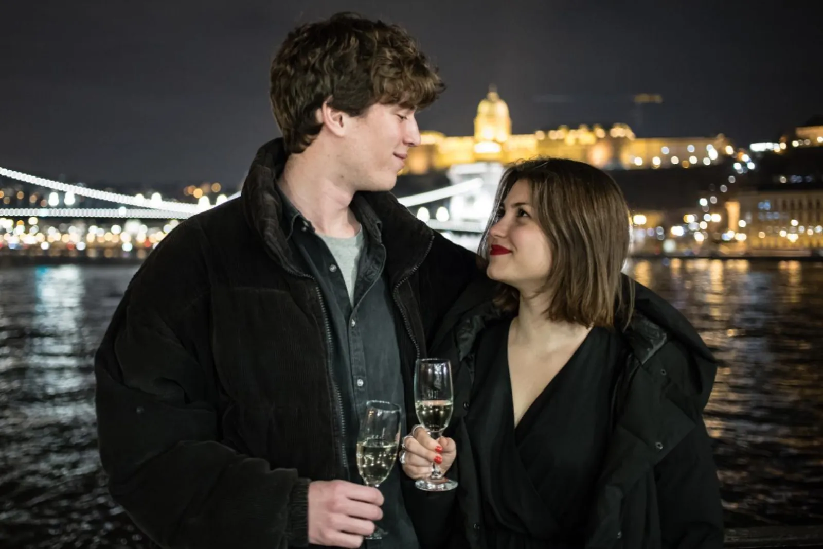 Couple holding sparkling wine on deck with lit Buda Castle across the Danube