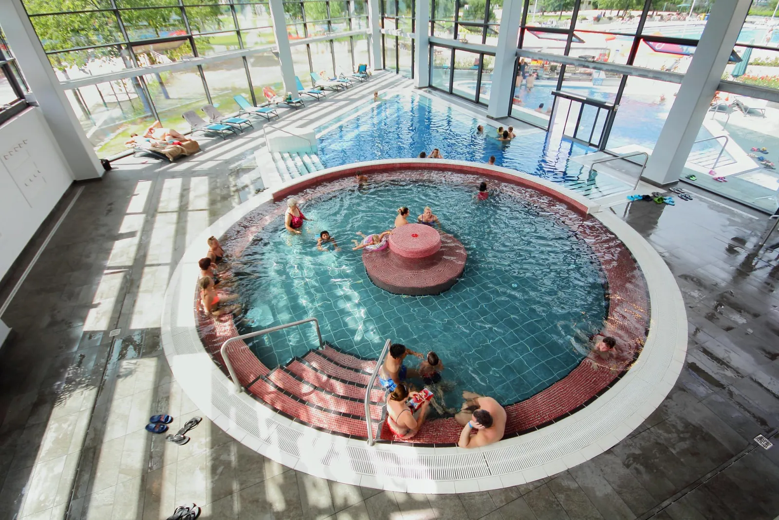 Round indoor thermal pool at Paskál seen from above, with the glass hall and outdoor pool beyond