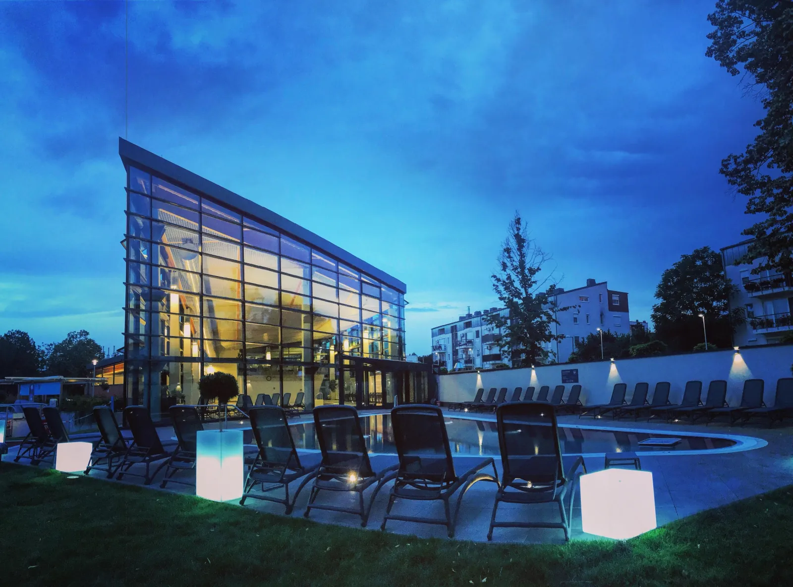 Paskál glass pool hall lit at dusk, with an outdoor pool, loungers and glowing light cubes