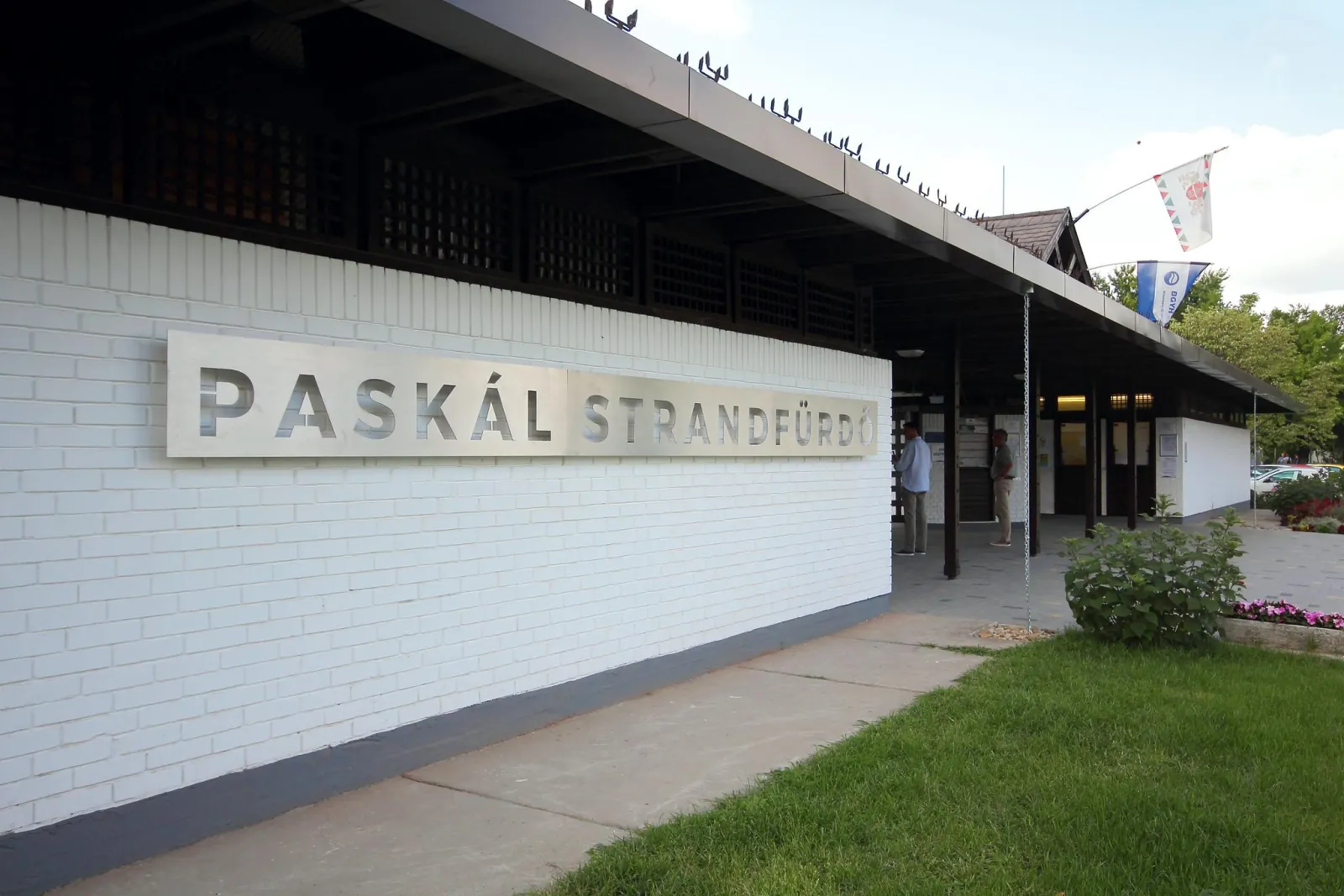 Paskál Strandfürdő entrance building with a white brick wall, lettering and flags