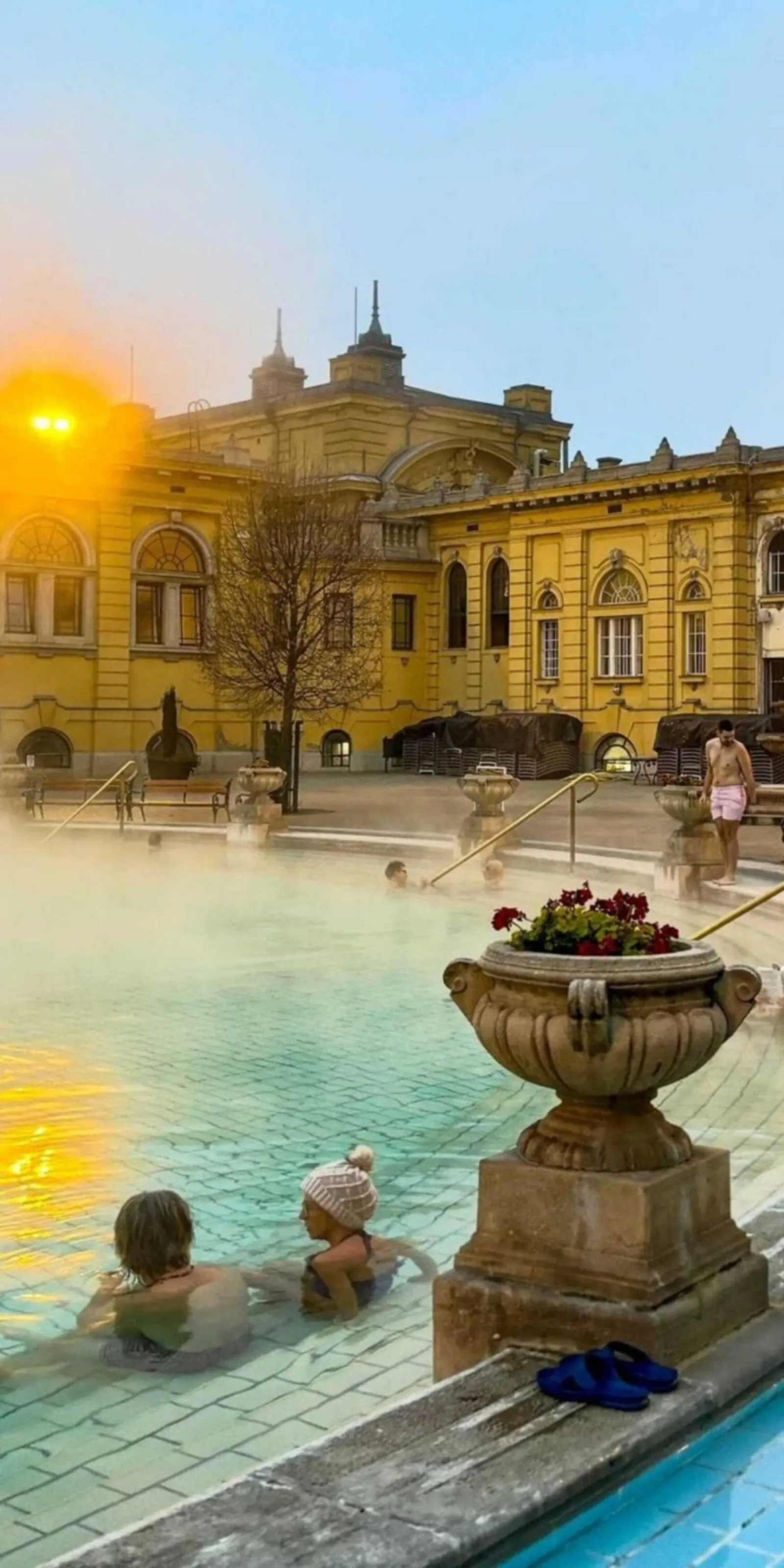 Bathers in a steaming Széchenyi outdoor pool beside a stone flower urn at sunrise