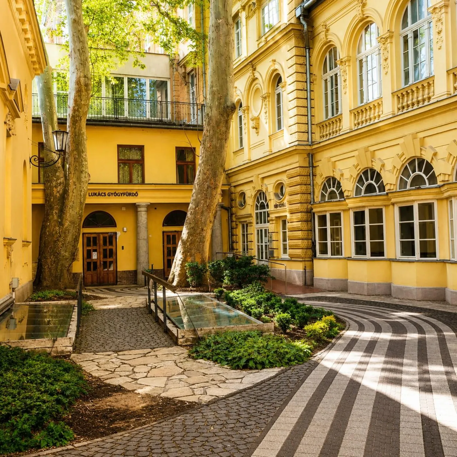 Lukács bath entrance courtyard with yellow facades, plane trees and striped paving