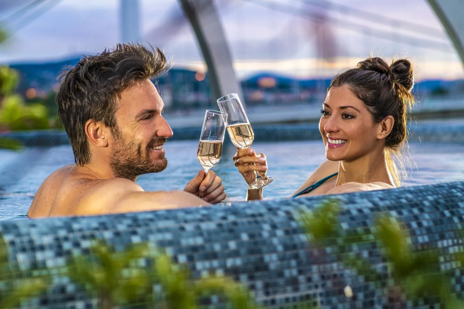 Couple toasting with sparkling wine in a mosaic-tiled pool above the river at dusk