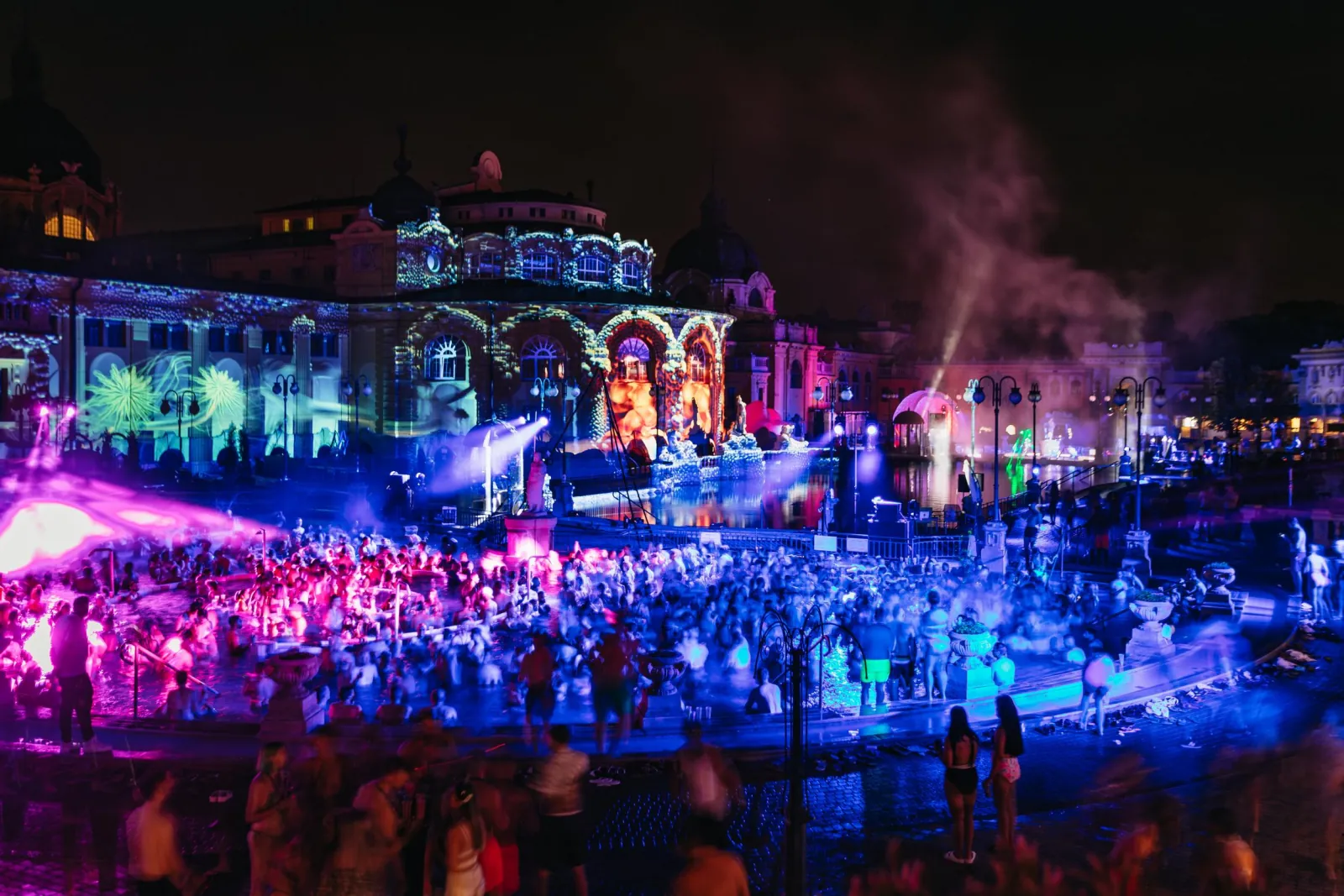 Packed Széchenyi outdoor pool under pink and blue party lights with projected facade