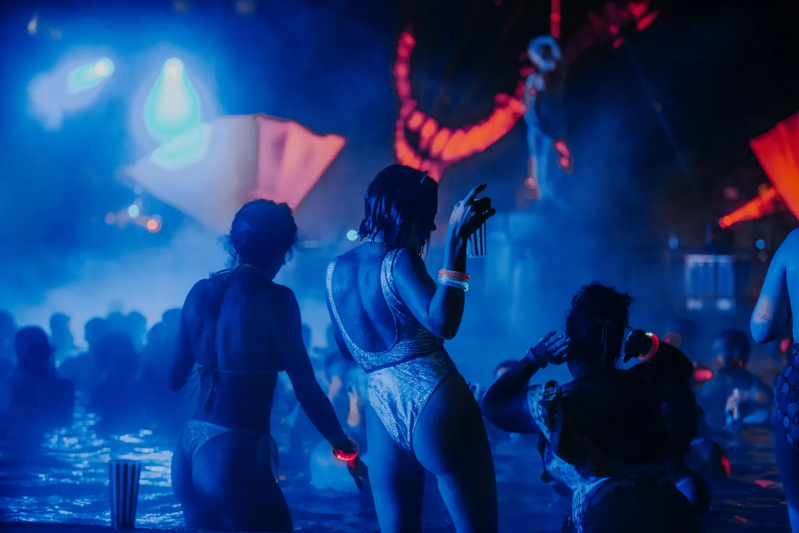 Partygoers in swimsuits dancing waist-deep in blue-lit water at Sparty