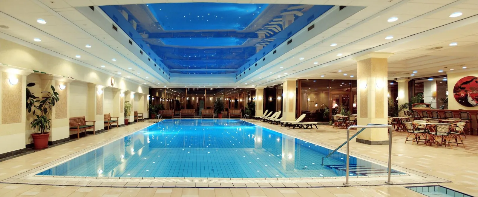 Long indoor swimming pool under a blue-lit ceiling with loungers and café tables