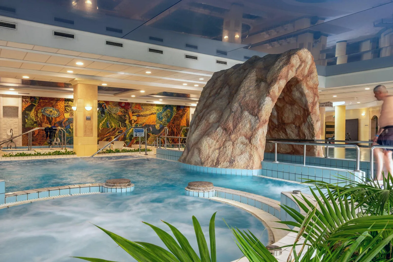 The Ensana Margaret Island thermal pool hall, with a rock grotto, palms and bathers in the warm water