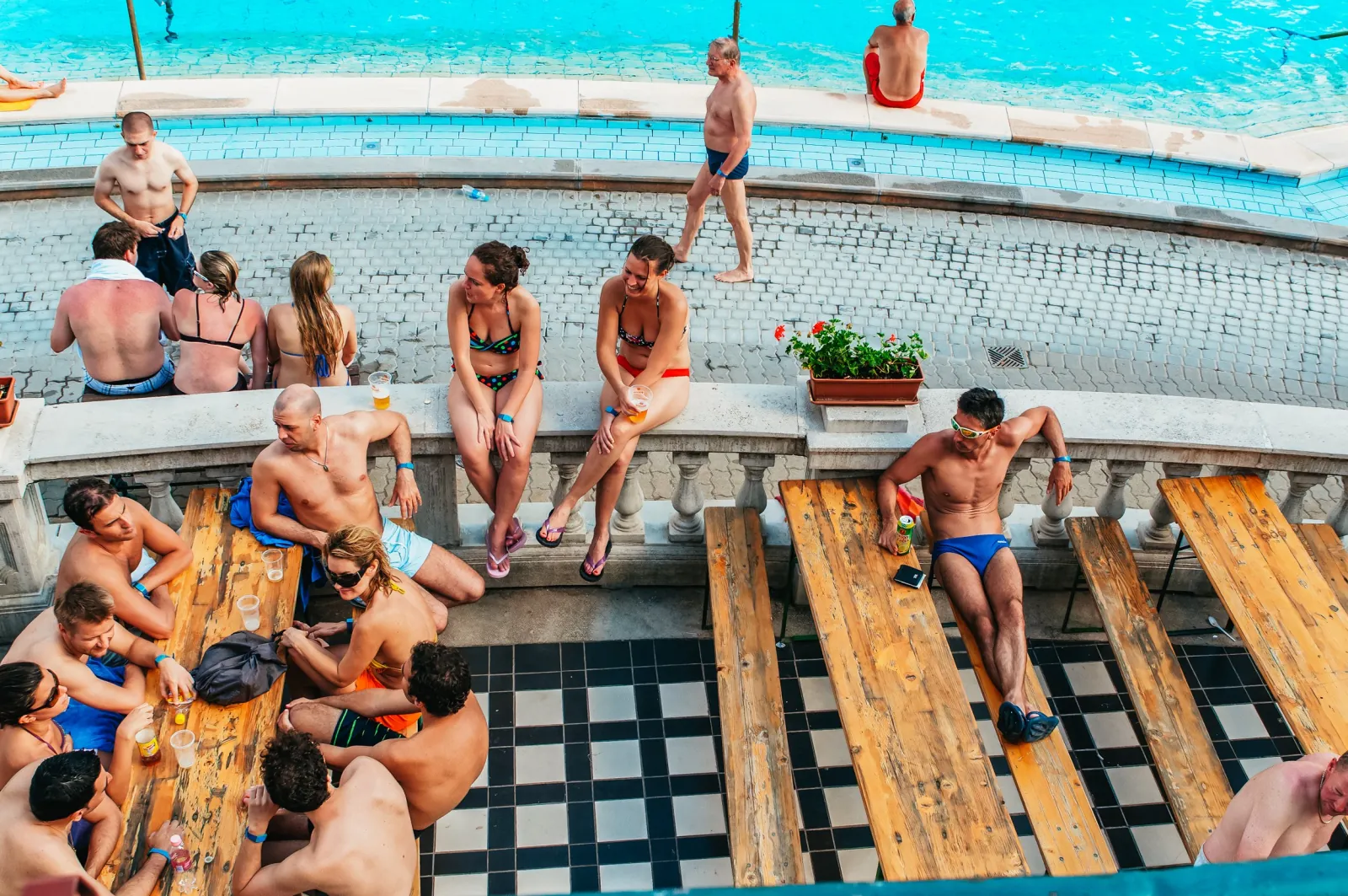 Bathers sitting on wooden benches and a balustrade beside a Széchenyi outdoor pool