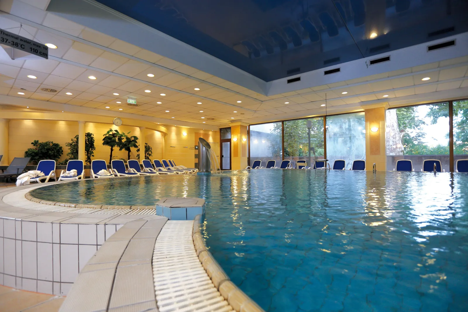 Curved edge of a large indoor pool with blue loungers and tall windows at the Ensana spa