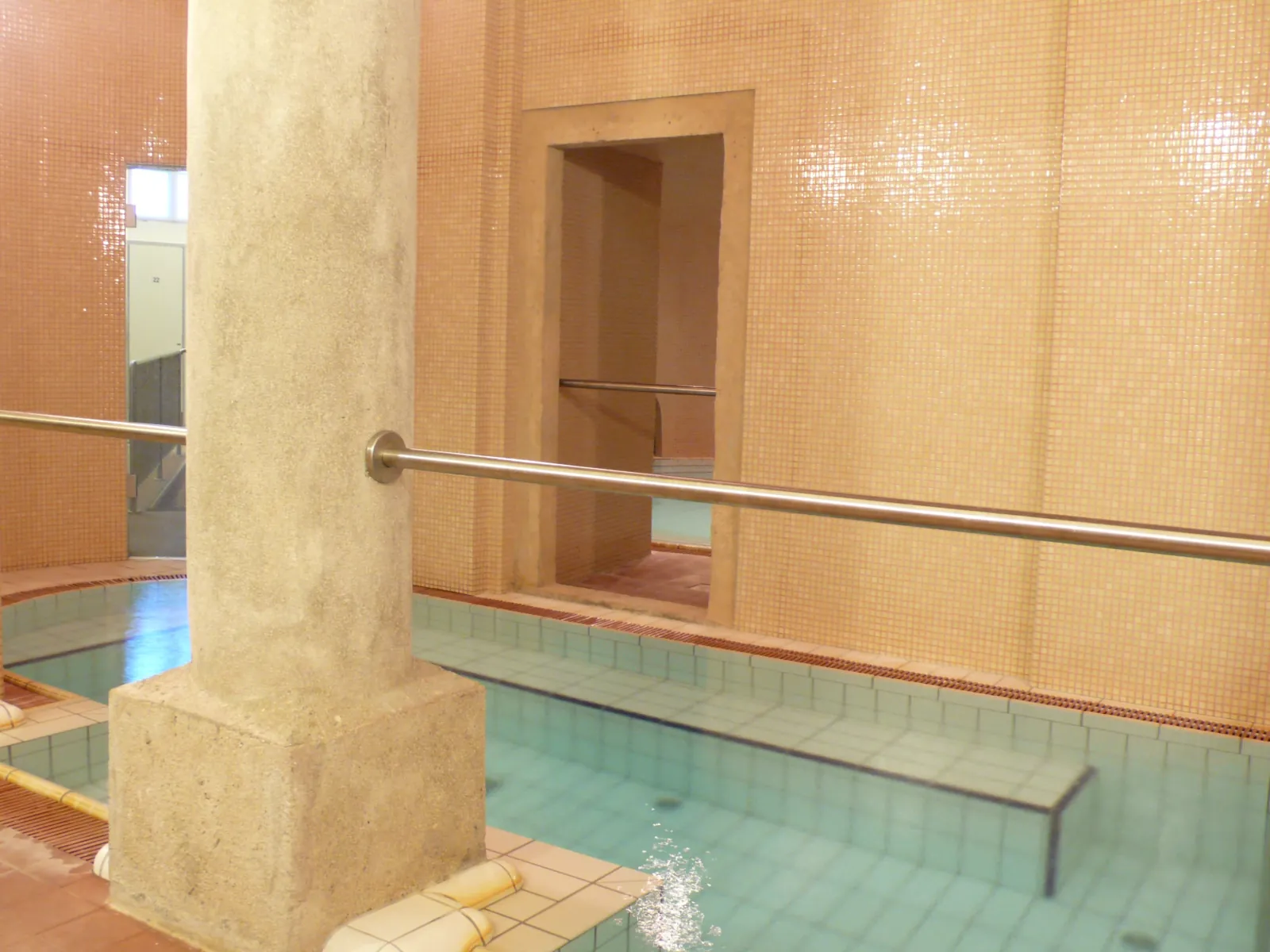 Indoor thermal pool at Lukács with peach mosaic walls, a stone pillar and a handrail