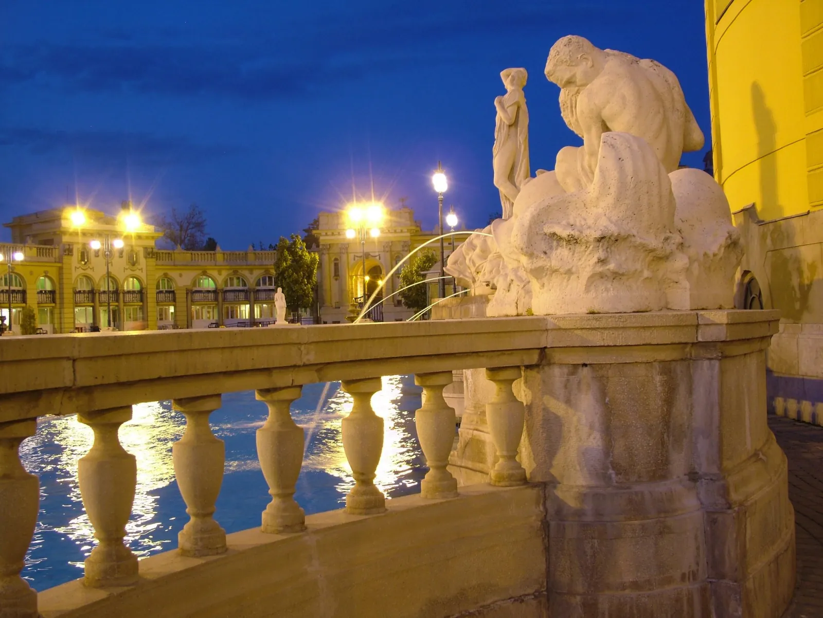 Stone statue and balustrade above Széchenyi's lit outdoor pool at dusk