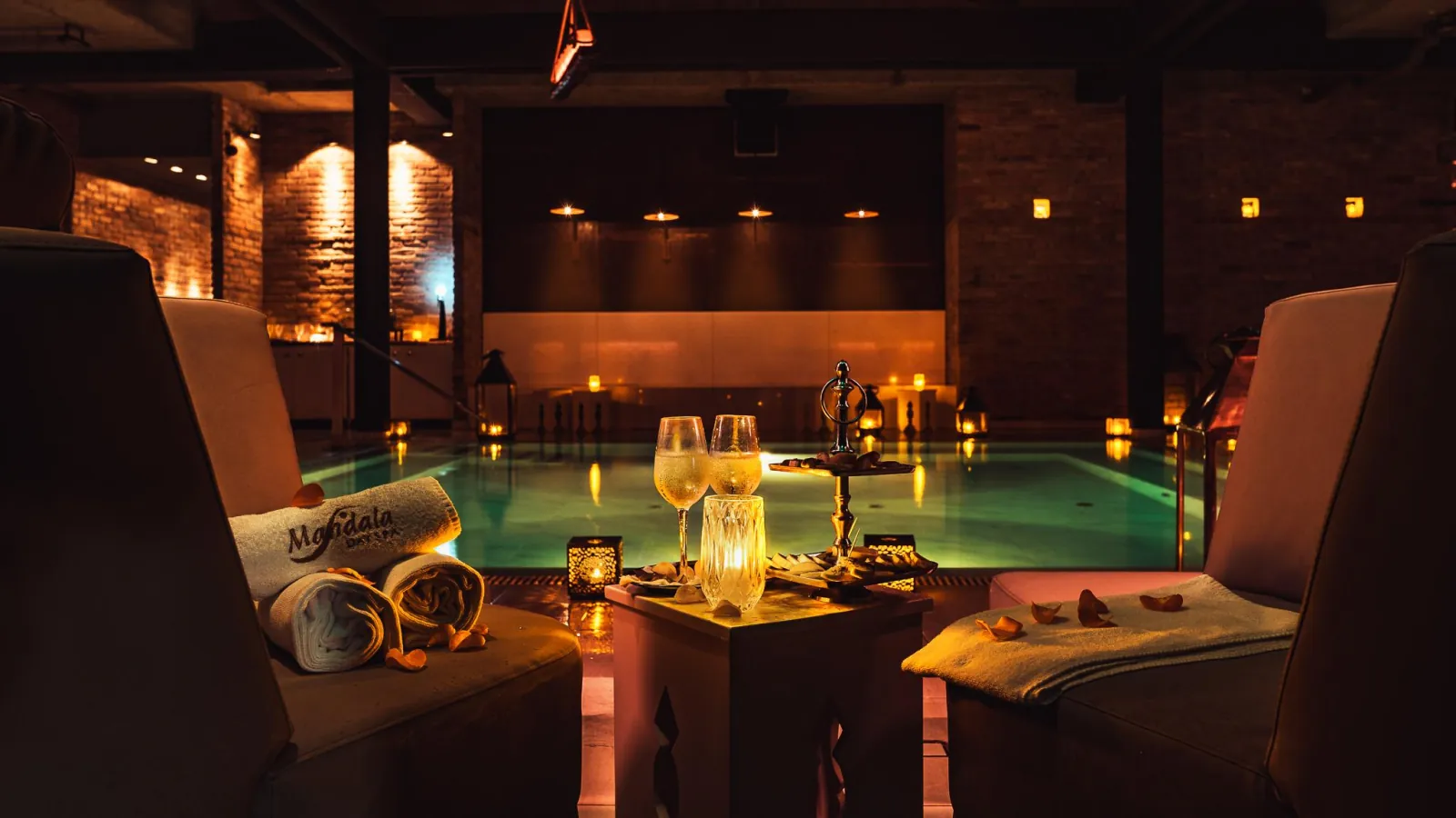Two armchairs with rolled Mandala towels and champagne glasses facing a candlelit pool