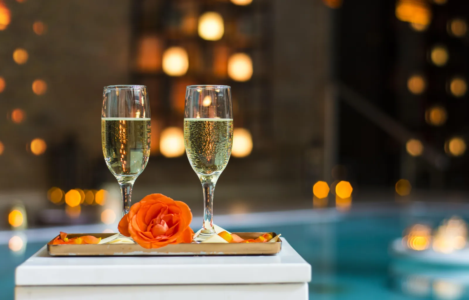Two glasses of champagne and an orange rose on a tray beside a lit pool