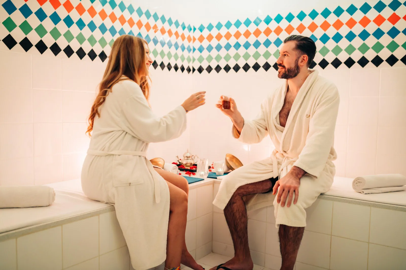 Couple in white robes sipping tea on a tiled bench under a diamond-patterned tile border