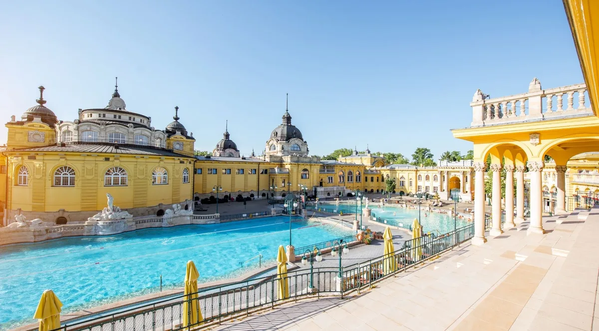 Széchenyi Thermal Bath's outdoor pools seen from the colonnaded terrace, the yellow neo-baroque domes against a clear blue sky