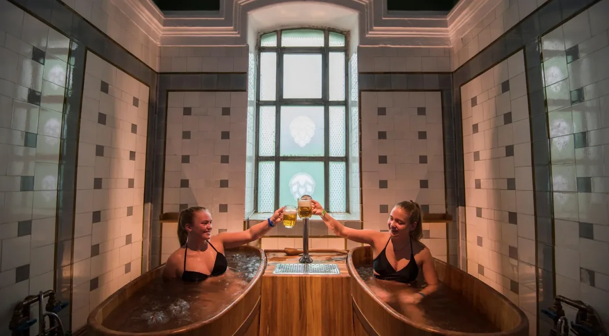 Two women toasting with mugs of beer from wooden tubs set side by side in a tiled room with a tall hop-patterned window