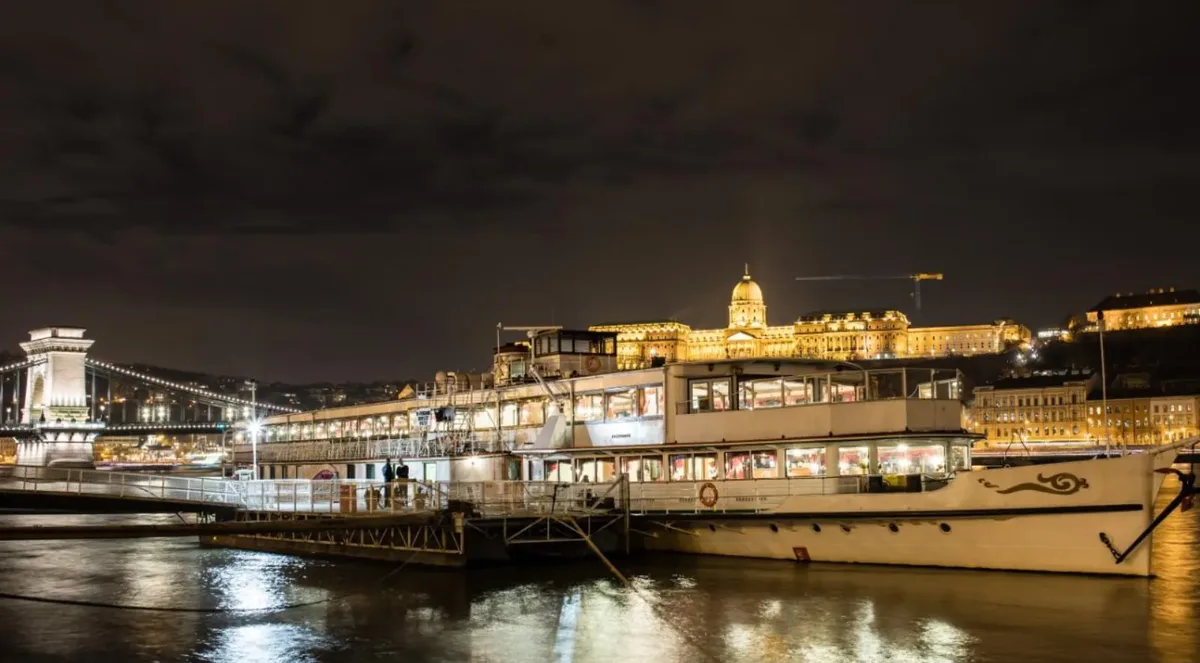 A white Danube sightseeing ship moored at night, with the floodlit Buda Castle and the Chain Bridge behind it