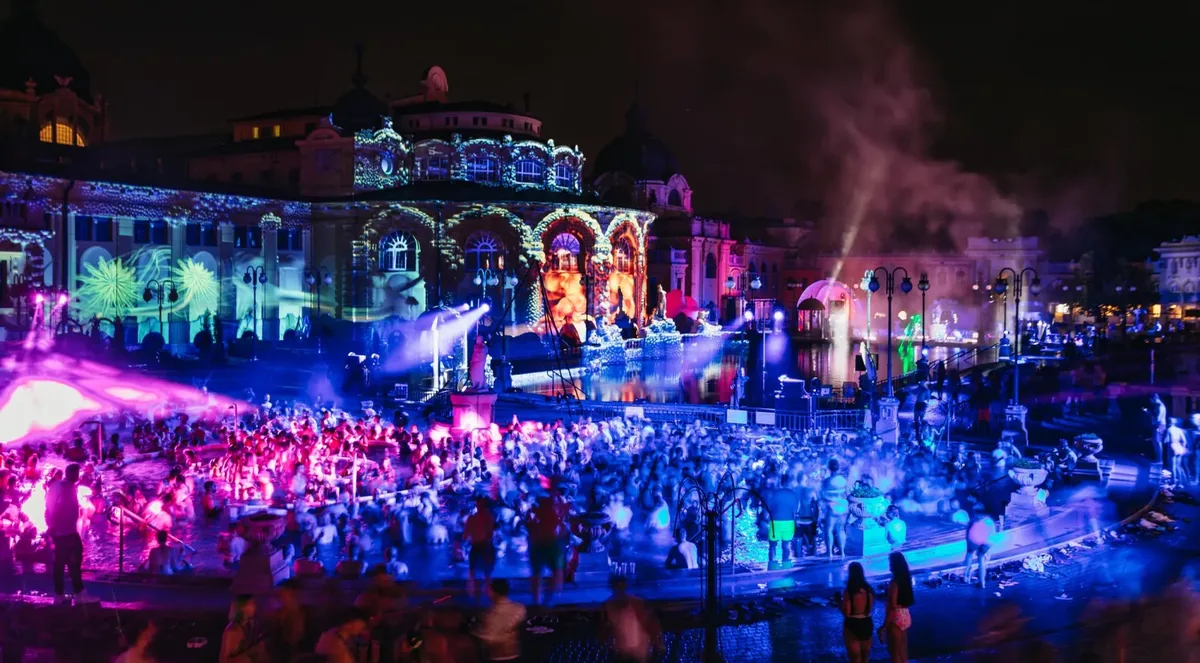 A crowd in the Széchenyi outdoor pool at night during Sparty, with the bathhouse facade lit by colored projections and stage lights