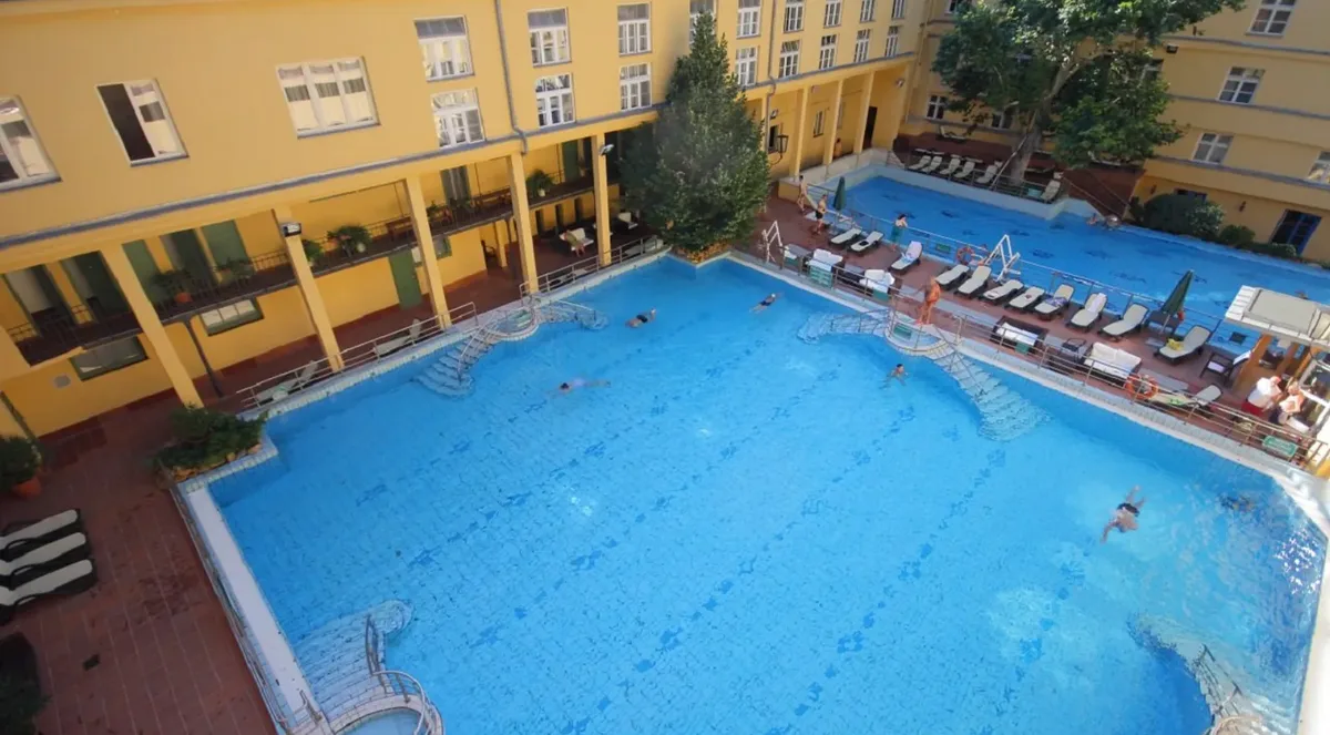 Swimmers in the large outdoor pool at Lukács Baths, seen from above, with yellow arcaded bathhouse wings and sun loungers around it