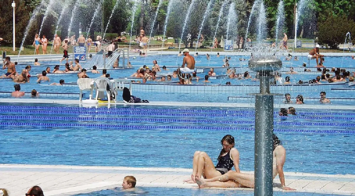 Bathers in the outdoor pools at Palatinus on a sunny summer day, with a row of water jets and trees behind the pool